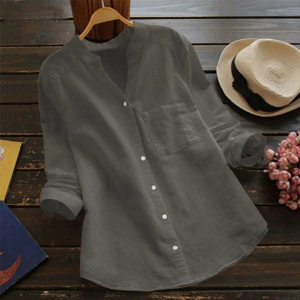 Graues Leinenhemd mit Stehkragen auf Holztisch, neben Strohhut und Blumen. Elegantes, lässiges Design für Damenmode.