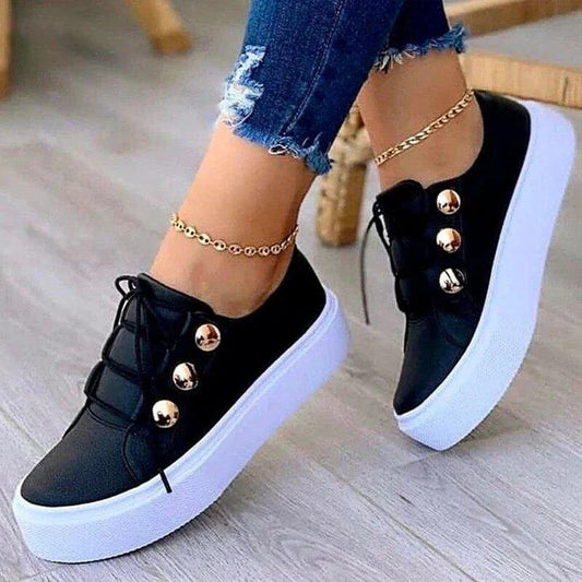 Schwarze Damen-Sneaker mit goldenen Knöpfen und weißer Plateau-Sohle, getragen mit zerrissenen Jeans und goldenen Fußkettchen.