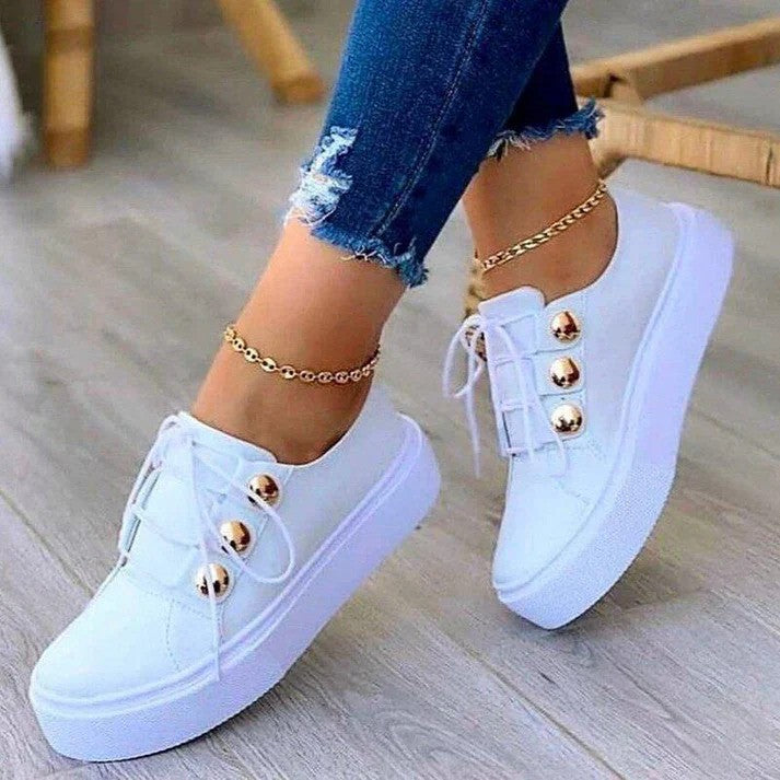 Weiße Damen-Sneaker mit goldenen Knöpfen, kombiniert mit blauen Jeans und Fußkettchen. Modische Freizeitschuhe für Damen.