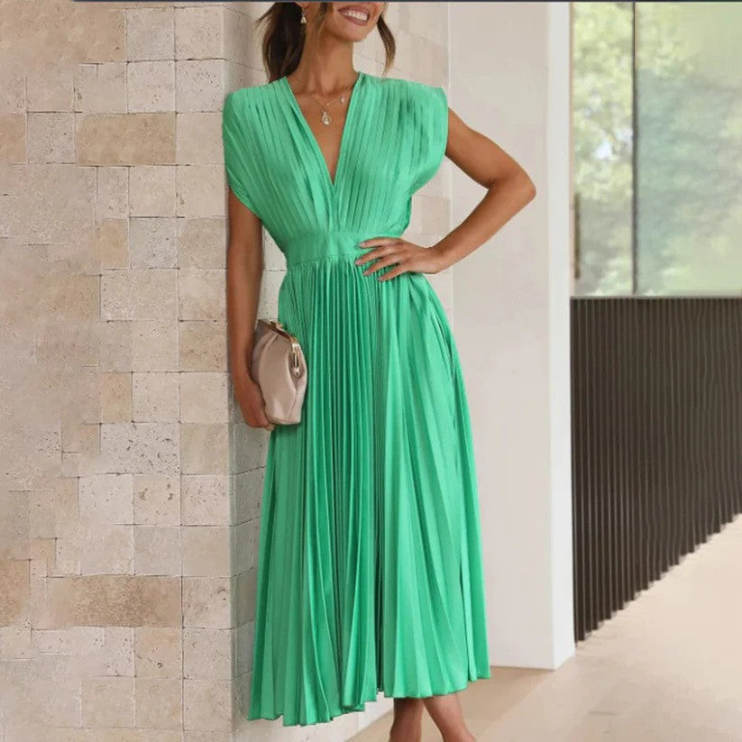 Frau in elegantem, grünem Plisseekleid lehnt an Steinwand, hält goldene Clutch. Stilvolle Abendmode, Sommerkleid, Modefotografie.