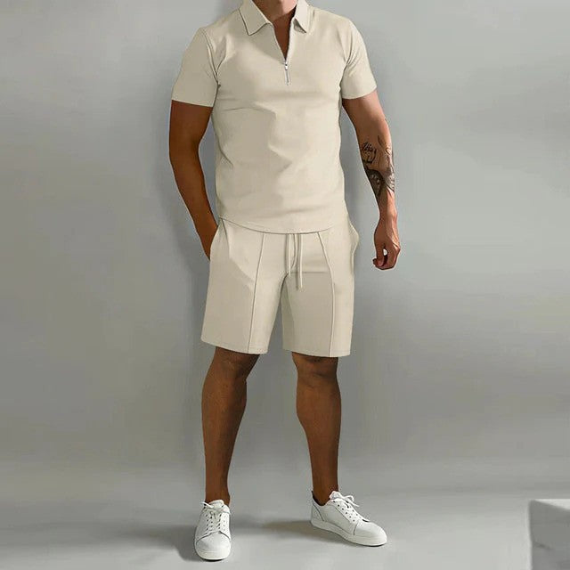 Mann in beigem Poloshirt und Shorts, tätowierter Arm, weiße Sneaker, grauer Hintergrund. Sommermode, Herrenoutfit, lässiger Stil.