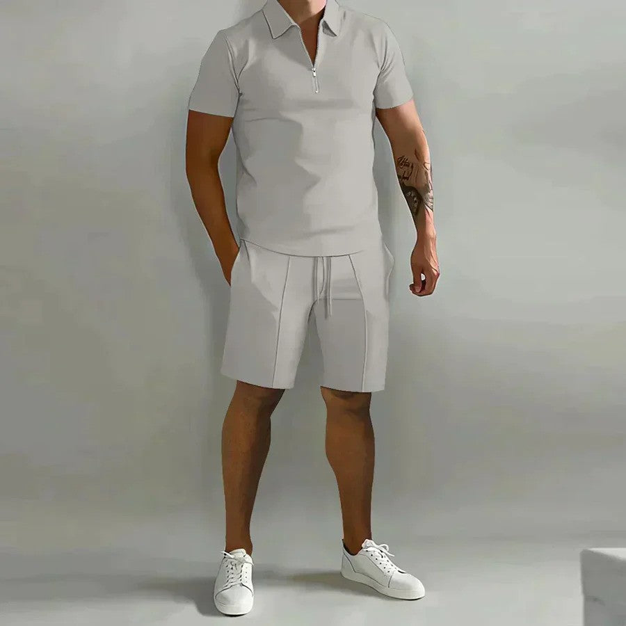 Mann in grauem Poloshirt und Shorts, weiße Sneaker, neutraler Hintergrund. Sommermode, Freizeitkleidung, Herrenoutfit, sportlich-elegant.