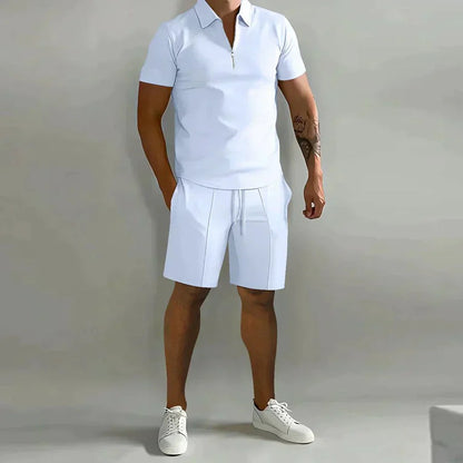 Mann in weißem Polohemd und Shorts, sportlich-eleganter Look, Sommermode, lässiger Stil, weiße Sneaker, grauer Hintergrund.