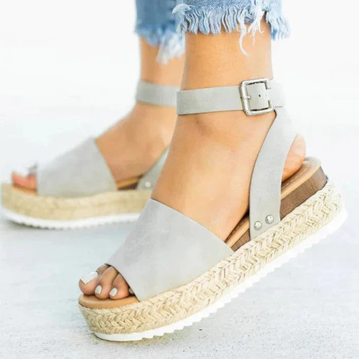 Graue Damen-Sandalen mit Keilabsatz und Knöchelriemen, kombiniert mit ausgefransten Jeans. Modische Sommerschuhe für Damen.