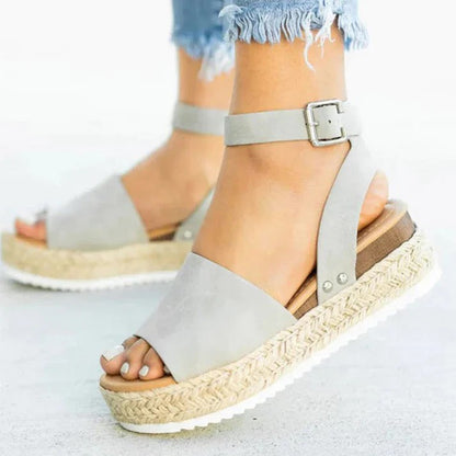 Graue Damen-Sandalen mit Keilabsatz und Knöchelriemen, kombiniert mit ausgefransten Jeans. Modische Sommerschuhe für Damen.