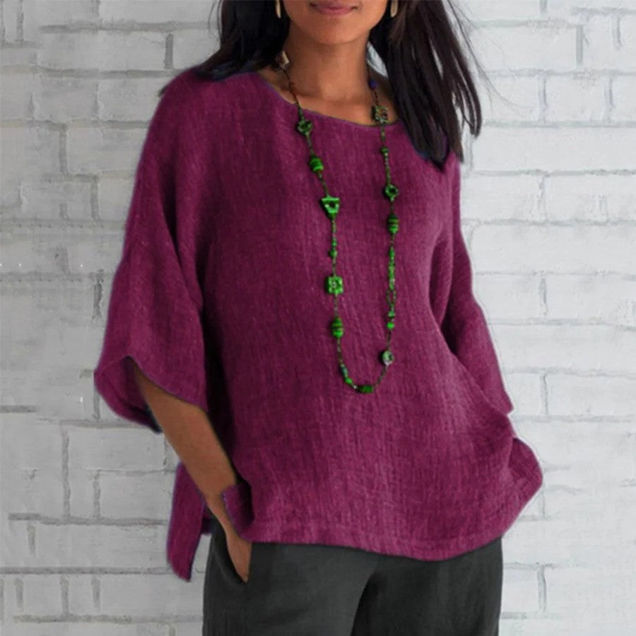 Frau in lila Leinenbluse vor weißer Ziegelwand, trägt lange grüne Halskette. Modetrend, lässiger Stil, Damenbekleidung, Herbstmode.