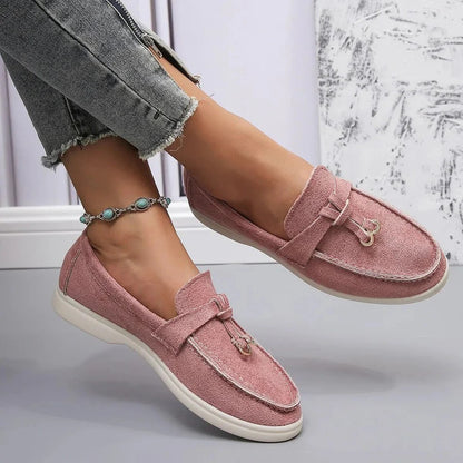 Damenfüße in rosa Mokassins, kombiniert mit grauen Jeans und einem türkisfarbenen Fußkettchen, vor einem modernen, minimalistischen Hintergrund.