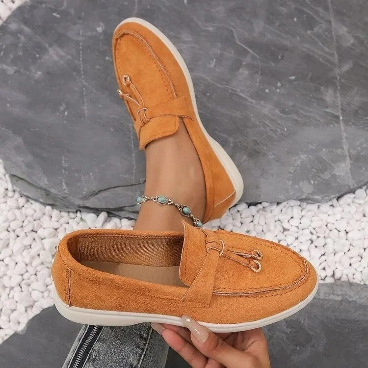 Orange Damen-Loafer aus Wildleder, getragen auf grauem Steinboden mit weißen Kieselsteinen. Modische Freizeitschuhe, ideal für den Sommer.
