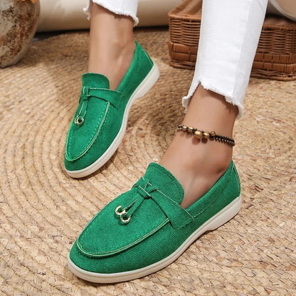 Grüne Damen-Loafer mit goldenen Details, getragen mit weißen Jeans. Modische, bequeme Schuhe auf geflochtenem Teppich. Ideal für Frühling und Sommer.