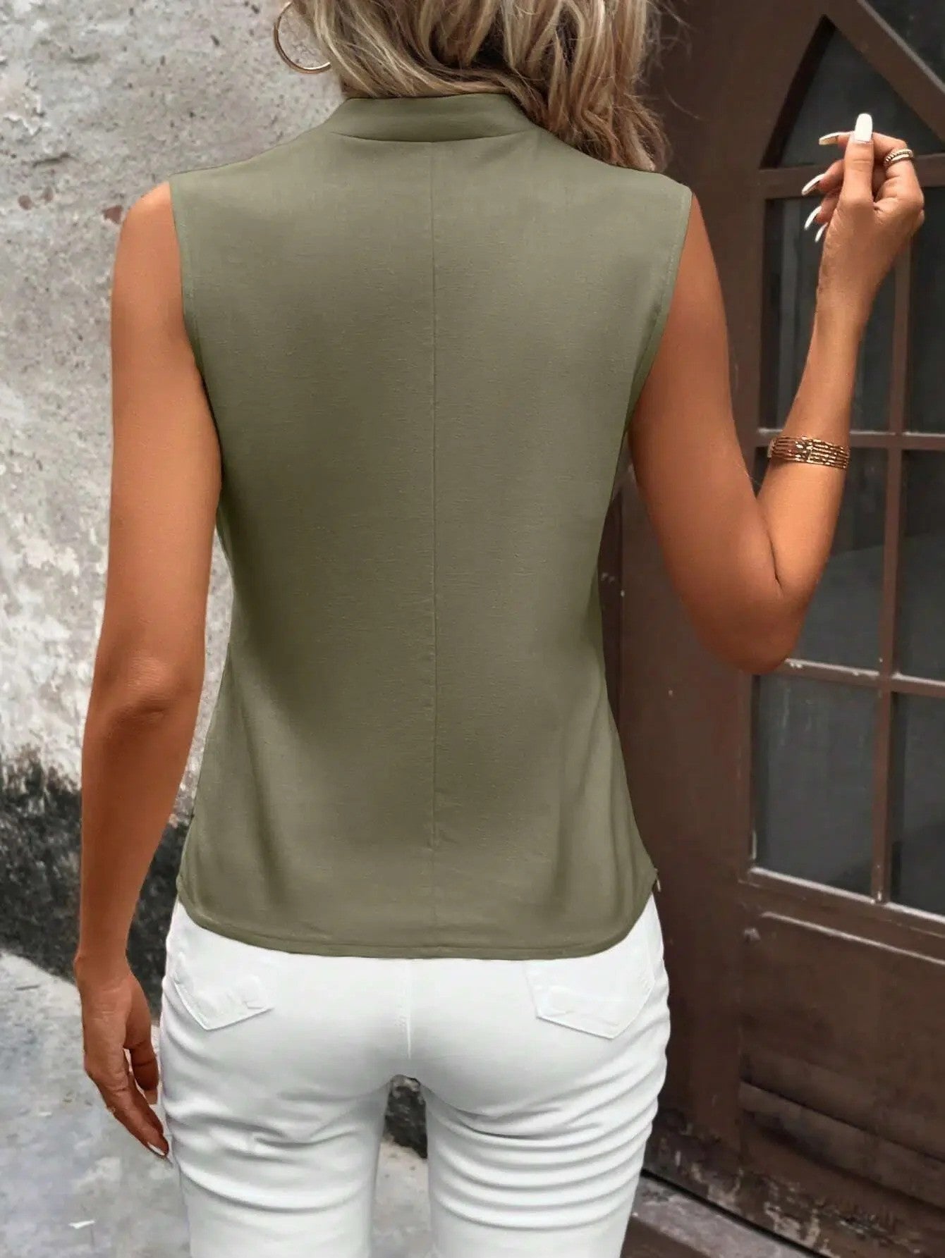 Frau in ärmellosem, olivgrünem Top und weißen Jeans vor rustikaler Wand. Sommermode, lässiger Stil, Rückansicht.