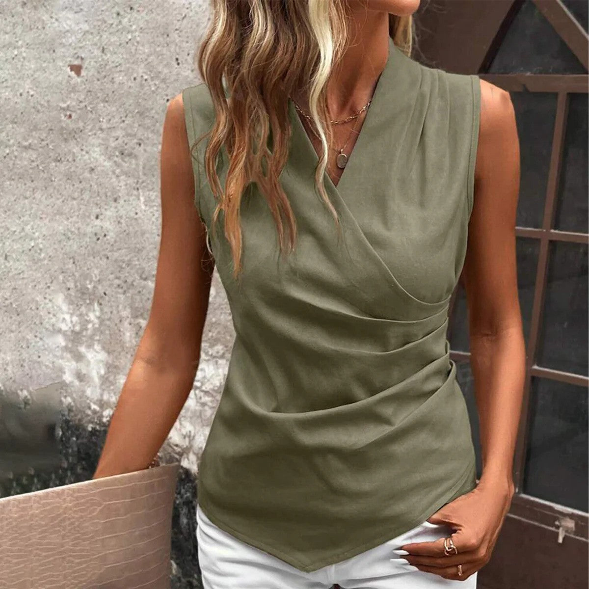Frau in ärmelloser, olivgrüner Bluse mit V-Ausschnitt, vor rustikaler Wand. Modetrend, Damenmode, Sommeroutfit, stilvolle Kleidung.