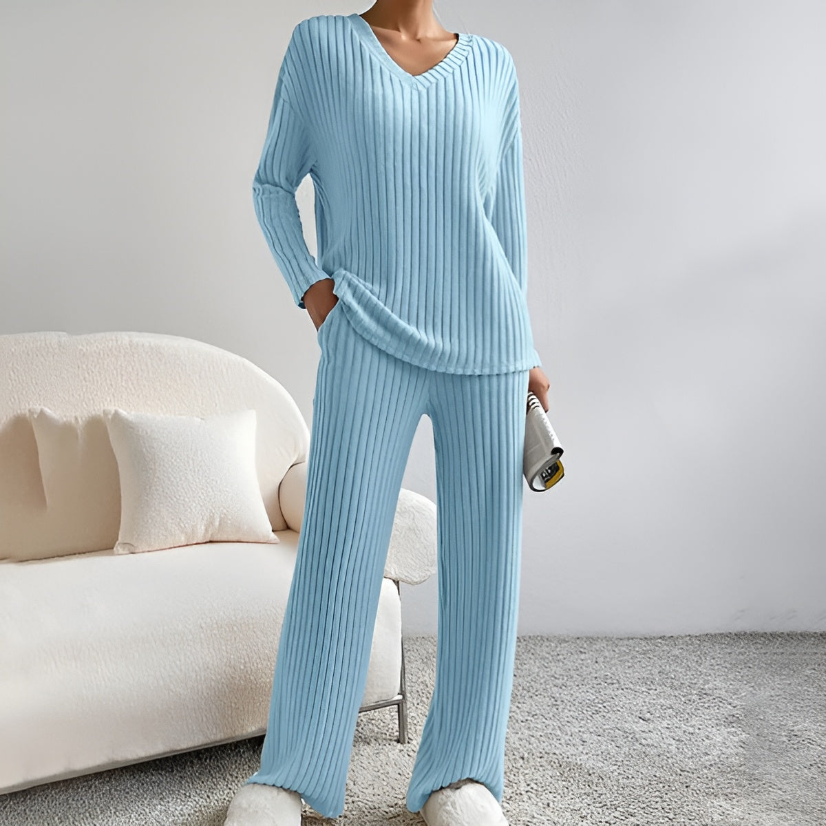 Frau in hellblauem, geripptem Lounge-Set mit V-Ausschnitt und flauschigen Hausschuhen, steht in modernem Wohnzimmer mit beigem Sofa und Kissen.