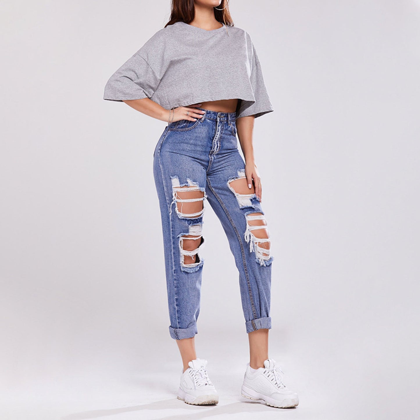 Frau in zerrissenen Jeans und grauem Crop-Top, trägt weiße Sneaker. Modetrend, lässiger Streetstyle, moderne Damenbekleidung.