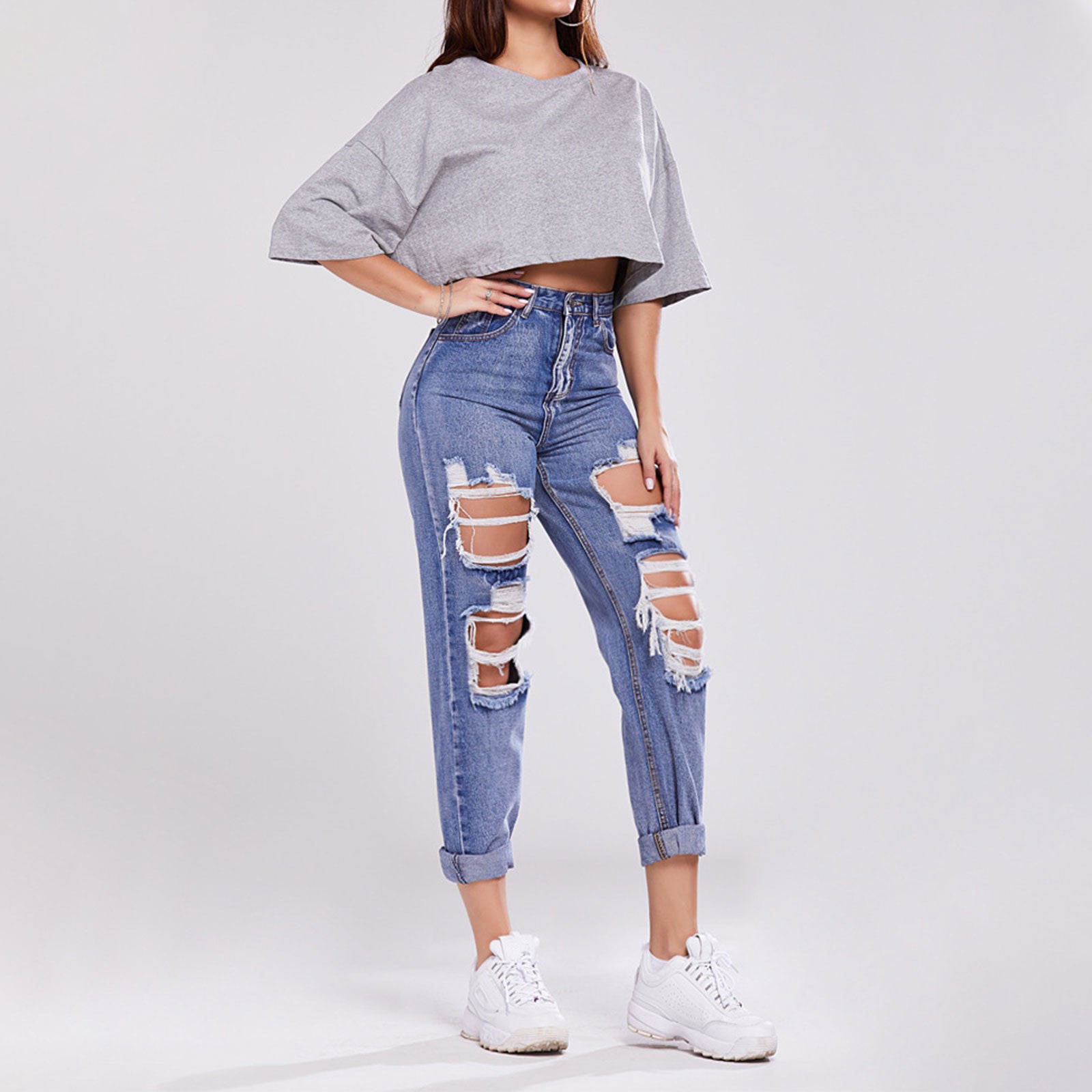 Frau in zerrissenen Jeans und grauem Crop-Top, trägt weiße Sneaker. Modetrend, lässiger Streetstyle, moderne Damenbekleidung.