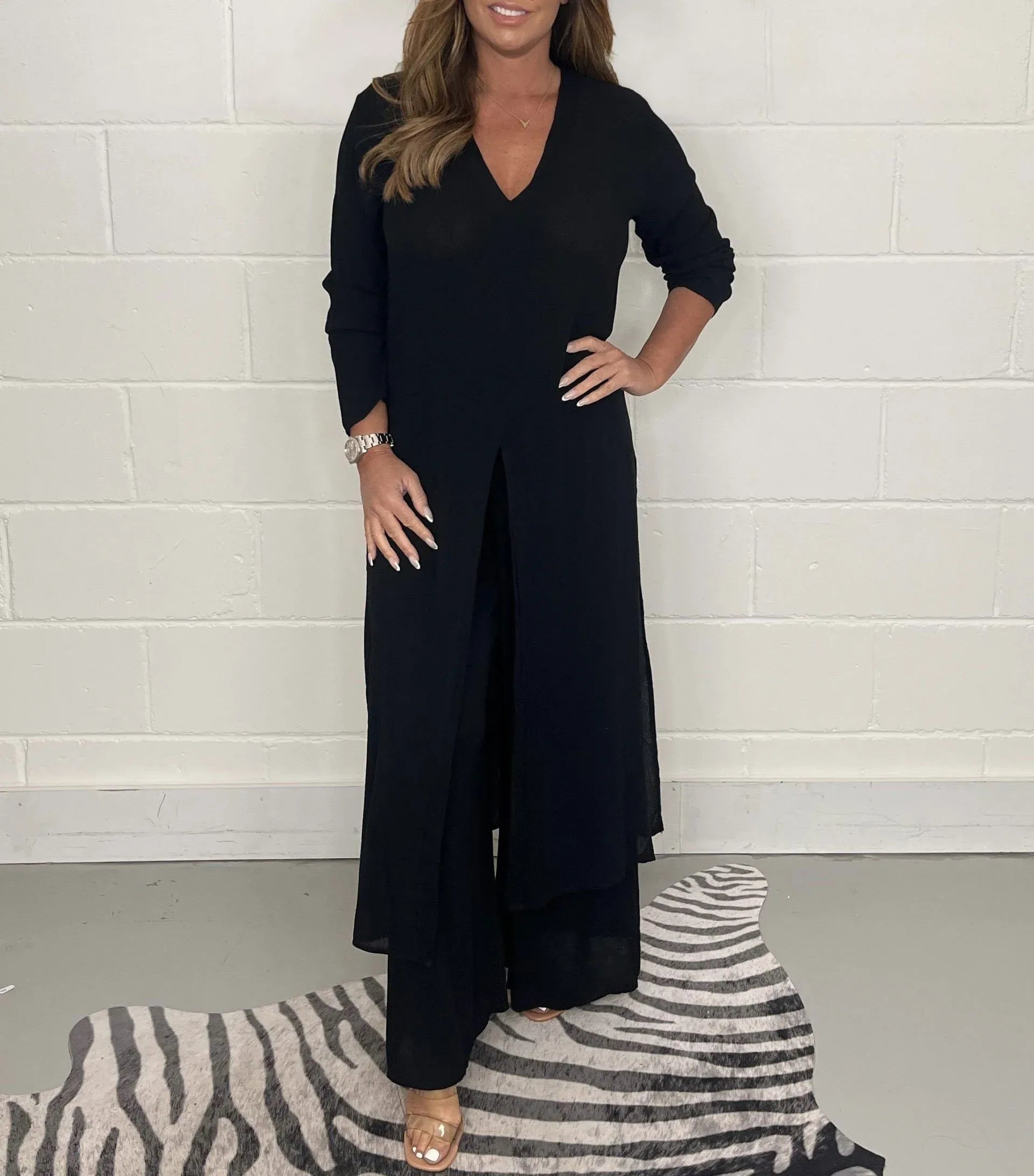 Frau in schwarzem, langem Kleid mit V-Ausschnitt, steht auf einem gestreiften Teppich vor einer weißen Wand. Elegante Mode, Abendkleid, Damenbekleidung.