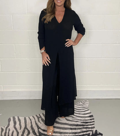 Frau in schwarzem, langem Kleid mit V-Ausschnitt, steht auf einem gestreiften Teppich vor einer weißen Wand. Elegante Mode, Abendkleid, Damenbekleidung.