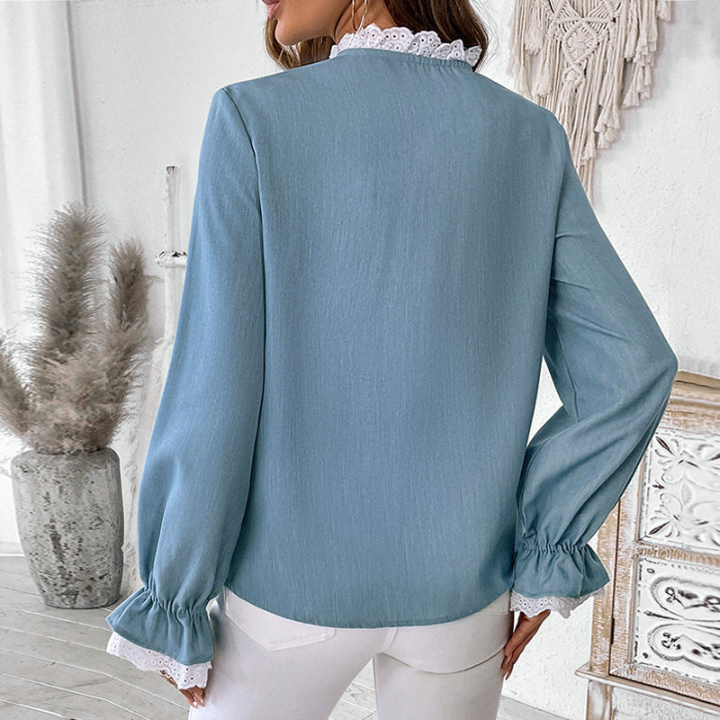 Frau in hellblauer Bluse mit Spitzenkragen und Rüschenärmeln, steht in modernem, hell eingerichtetem Raum. Elegante Damenmode, stilvoller Look.