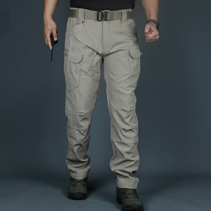 Mann in taktischer Hose, beige, mit Taschen und Gürtel, hält Funkgerät. Outdoor-Kleidung, robust, ideal für Wandern und Abenteuer.