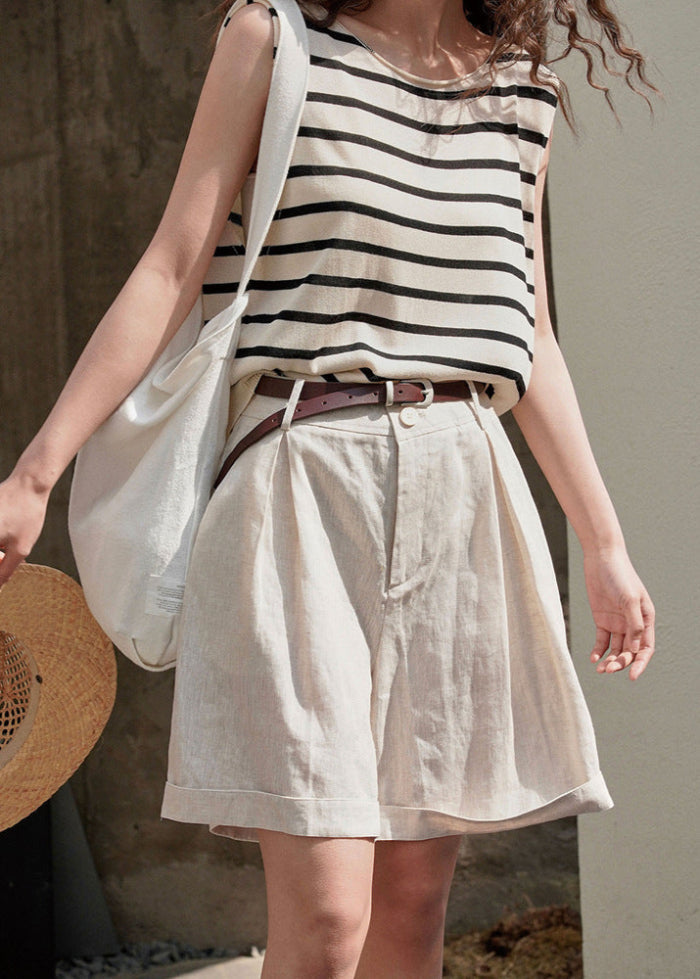 Frau in gestreiftem ärmellosen Top und beigen Shorts, trägt weißen Stoffbeutel und Strohhut. Sommermode, lässiger Stil, urbane Kleidung.