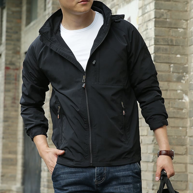 Mann in schwarzer Windjacke und blauer Jeans vor Backsteinwand. Modische Herrenbekleidung, lässiger Stil, Outdoor-Jacke.