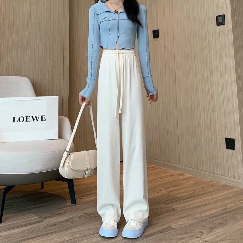 Frau in blauem Oberteil und weißen Hosen posiert in modernem Raum mit Loewe-Tasche. Modetrend, stilvolle Damenbekleidung, elegantes Outfit.