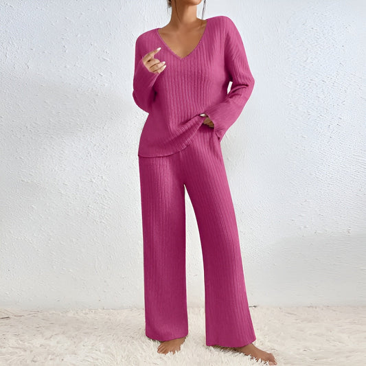 Frau in pinkem Strick-Zweiteiler, bestehend aus Pullover und Hose, steht barfuß auf weißem Teppich vor weißer Wand. Modetrend, gemütlich, stilvoll.