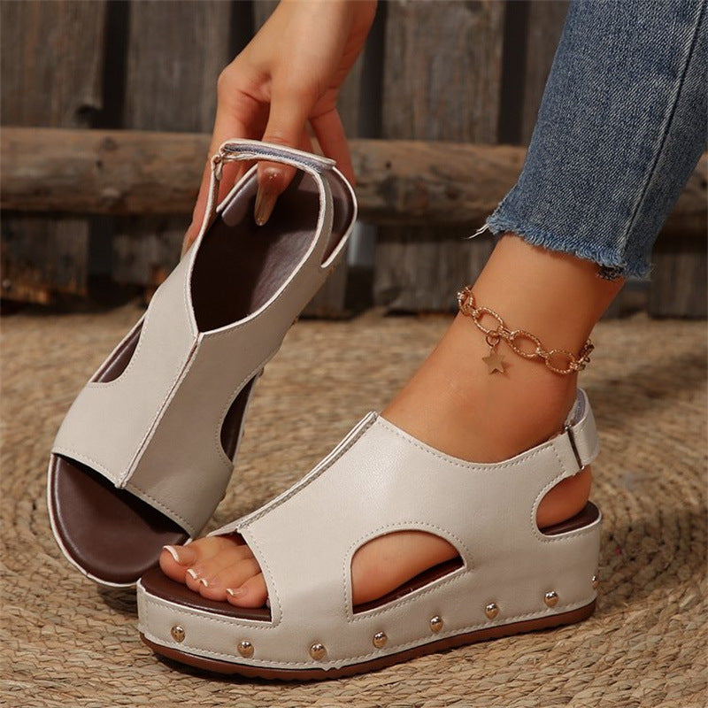 Beige Sandalen mit Keilabsatz und Nieten, getragen von einer Person mit Jeans und einem Stern-Armband. Modische Sommerschuhe für Damen.