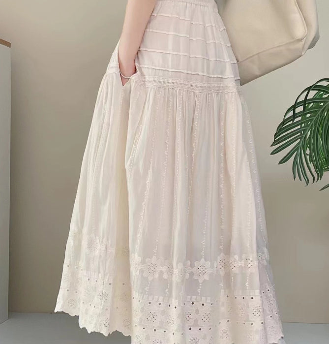 Frau in weißem, langem Spitzenrock mit Tasche, steht neben grüner Pflanze. Sommermode, eleganter Stil, luftiges Design.