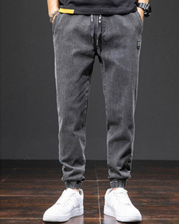 Graue Jogginghose für Herren, lässig und bequem, mit elastischem Bund und Bündchen, kombiniert mit weißen Sneakers auf dunklem Holzboden.