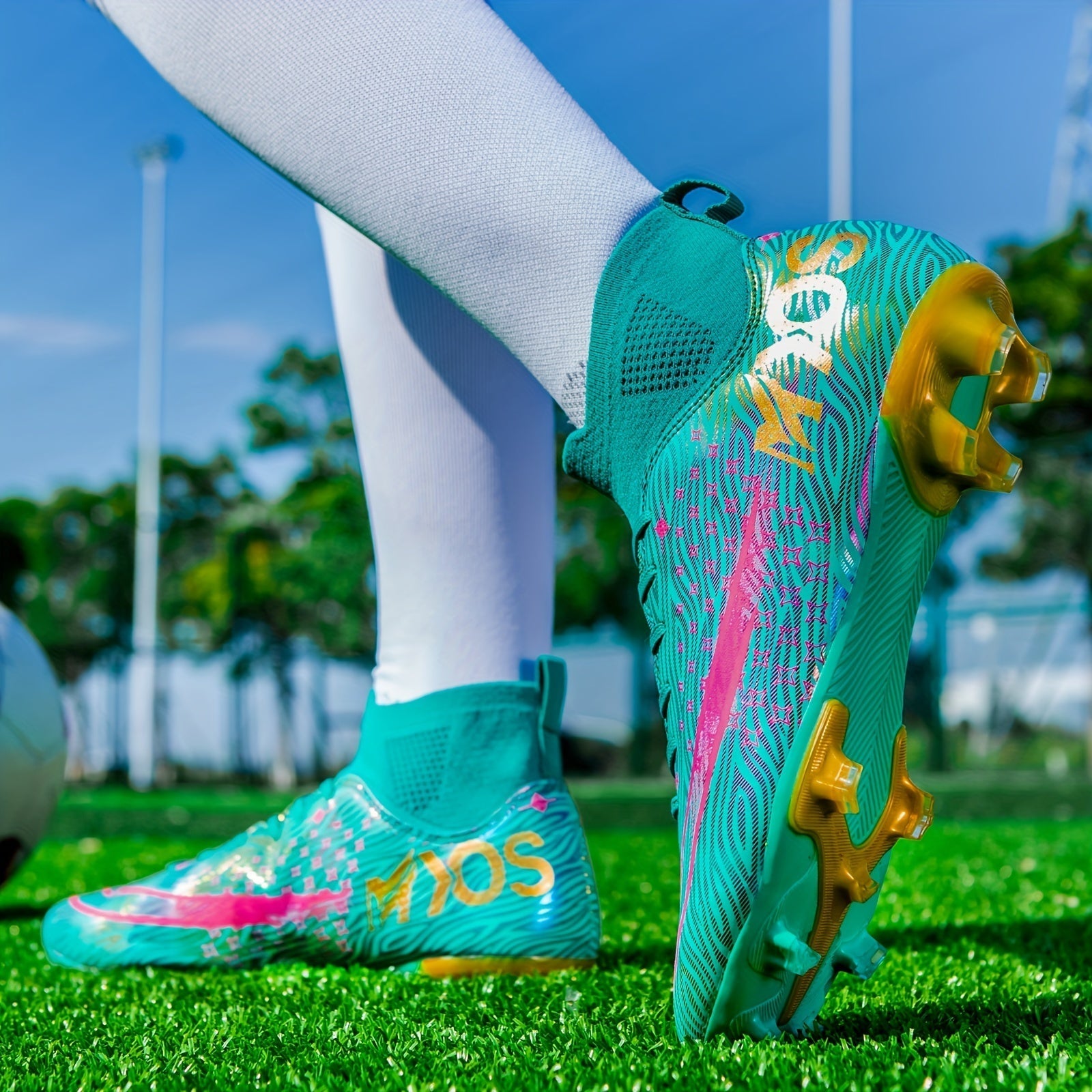 Fußballschuhe in Türkis mit pinken und goldenen Akzenten auf grünem Kunstrasen, getragen von Person in weißen Socken, Fußball im Hintergrund.