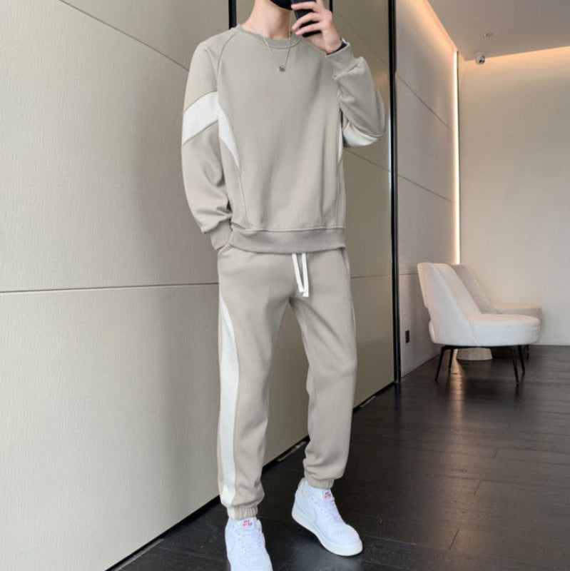 Person in beigem Trainingsanzug mit weißen Streifen, steht in modernem Raum. Weiße Sneakers, minimalistische Einrichtung, modischer Freizeitlook.