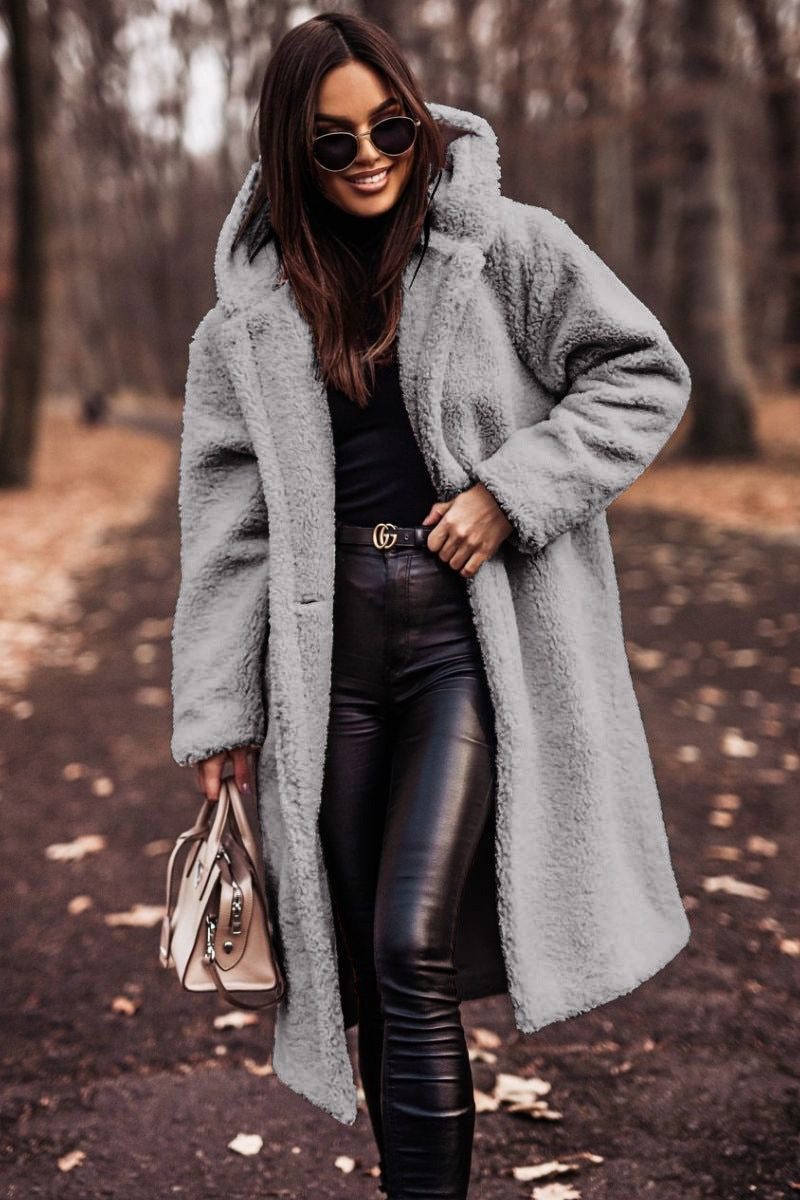 Frau im grauen Teddy-Mantel, schwarze Lederhose, Sonnenbrille, hält beige Handtasche, Herbstwald. Modetrend, Herbstmode, stilvoll gekleidet.