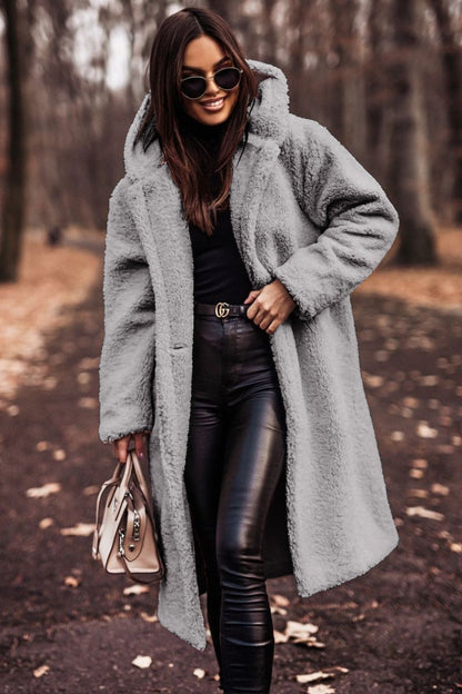 Frau im grauen Teddy-Mantel, schwarze Lederhose, Sonnenbrille, hält beige Handtasche, Herbstwald. Modetrend, Herbstmode, stilvoll gekleidet.