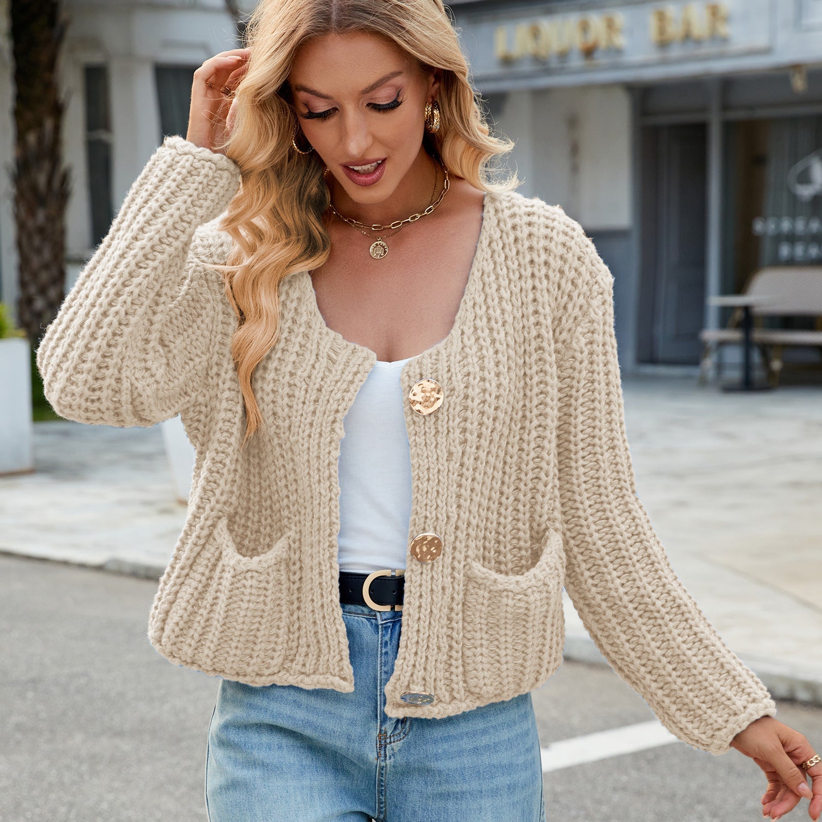 Frau trägt beige Strickjacke mit Knöpfen und Taschen, kombiniert mit weißem Top und blauer Jeans. Modetrend, Herbstmode, lässiger Stil.
