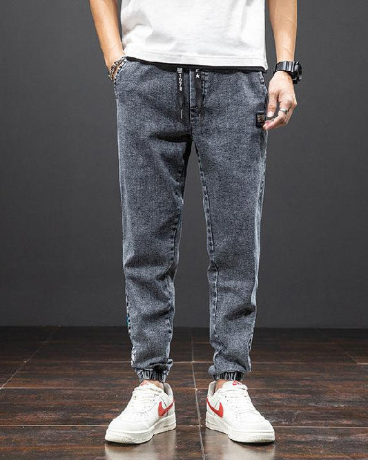 Mann trägt graue Jogginghose und weiße Sneakers mit rotem Logo auf dunklem Holzboden. Lässige Herrenmode, Streetwear, bequeme Freizeitkleidung.