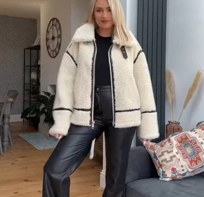 Frau in modischer Winterjacke aus Teddyfell und schwarzer Lederhose steht in modernem Wohnzimmer. Stilvolle Wintermode, gemütliches Interieur.
