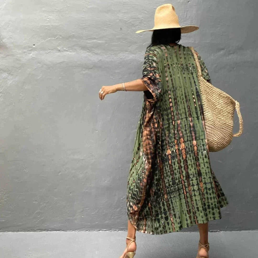 Frau in grünem Kaftan mit Muster, Strohhut und Strohtasche, geht auf grauem Hintergrund. Sommermode, Boho-Stil, lässiger Look.
