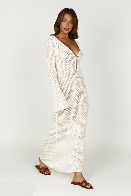 Frau in weißem, gehäkeltem Maxikleid mit tiefem Ausschnitt und langen Ärmeln, trägt braune Sandalen. Elegante Sommermode, Boho-Stil.