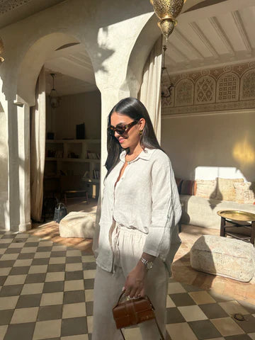 Frau in heller Kleidung und Sonnenbrille steht in elegantem, marokkanischem Innenraum mit Mosaikfliesen und traditionellen Lampen, hält eine braune Tasche.