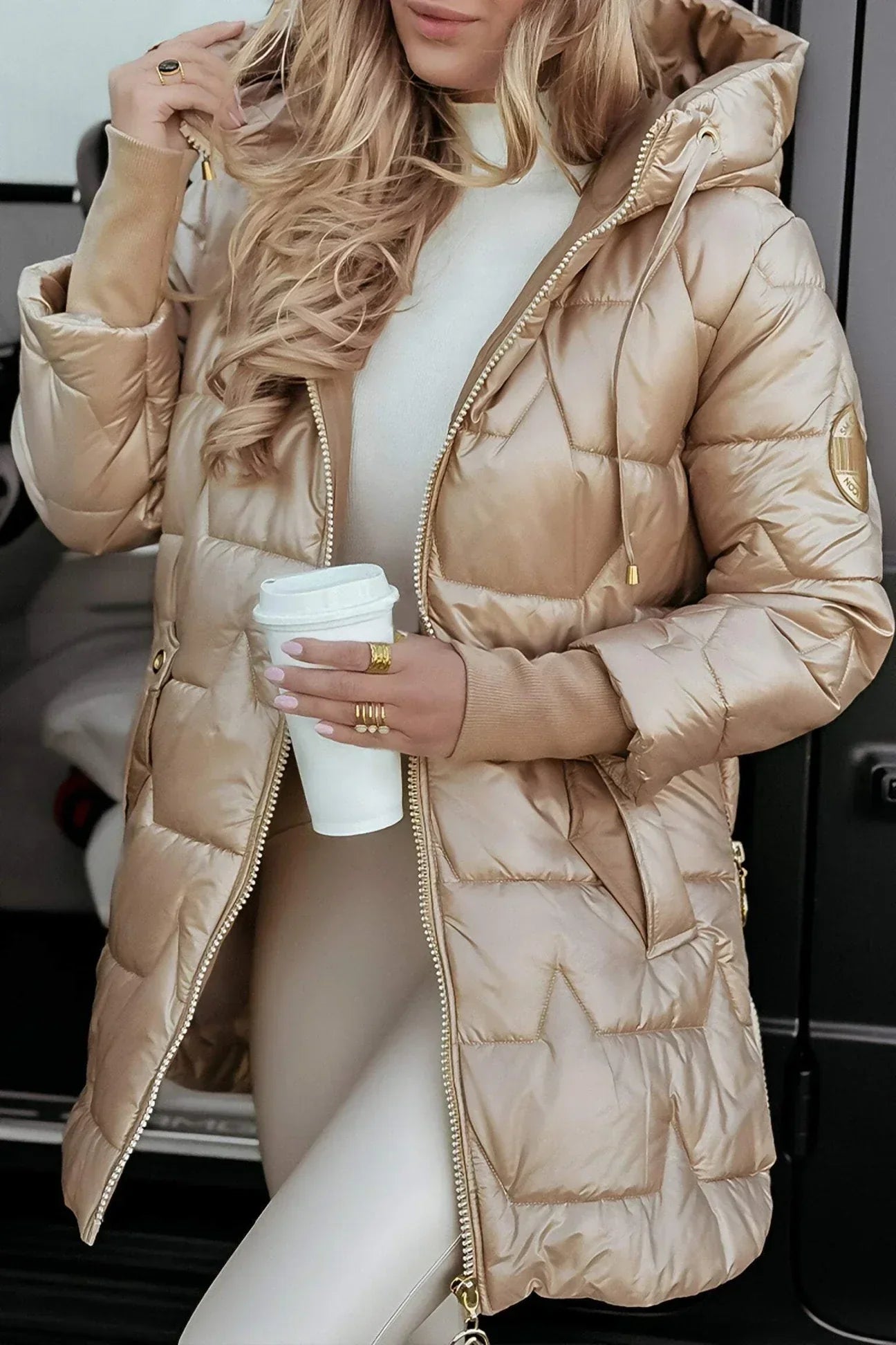Frau in beiger Daunenjacke mit Kapuze hält Kaffeebecher, modische Winterkleidung, eleganter Look, Outdoor, warme Jacke, Damenmode.