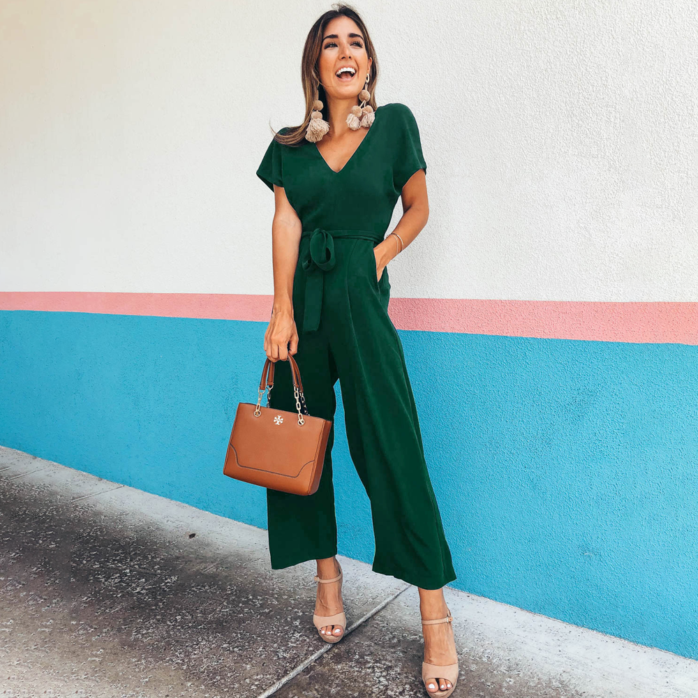 Frau in grünem Jumpsuit vor bunter Wand, hält braune Handtasche. Modetrend, Sommeroutfit, stilvolle Damenmode, elegante Accessoires.