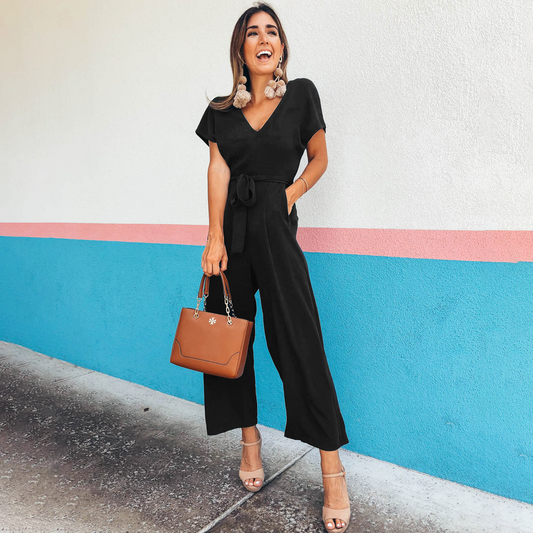 Frau in schwarzem Jumpsuit mit brauner Handtasche vor blau-rosa Wand. Modetrend, eleganter Look, Sommeroutfit, Damenmode, Streetstyle.