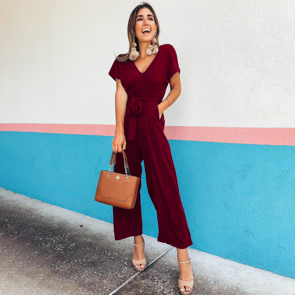Frau in rotem Jumpsuit vor bunter Wand, hält braune Handtasche. Modetrend, Sommeroutfit, elegante Damenmode, stilvolle Accessoires.
