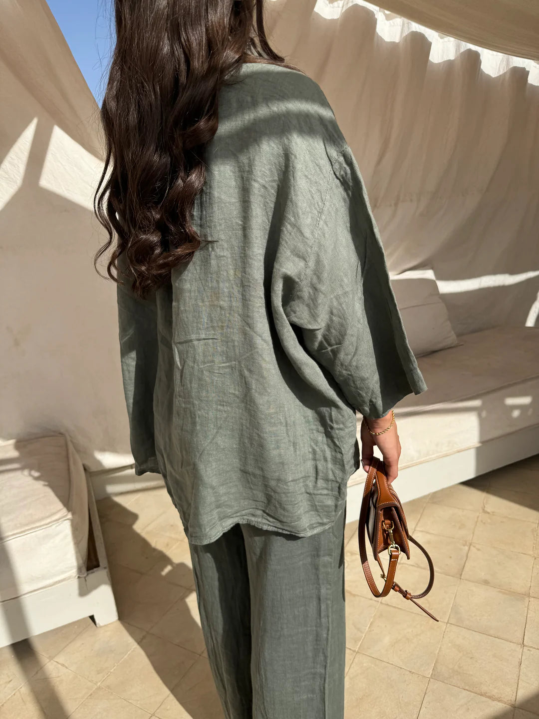Frau in grauem Leinenanzug mit brauner Handtasche, steht auf sonniger Terrasse. Sommermode, lässiger Stil, natürliche Materialien.
