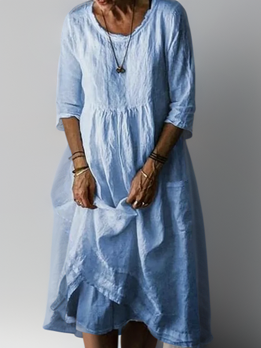 Frau in hellblauem Leinenkleid mit Dreiviertelärmeln, lässiger Stil, trägt Armbänder und Halskette, vor grauem Hintergrund.
