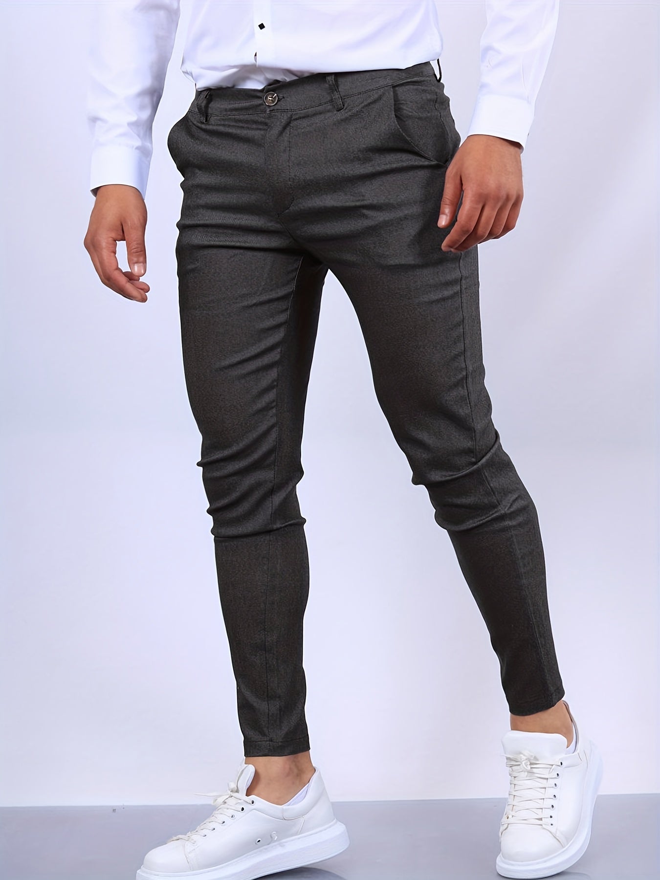 Mann in eleganter, dunkelgrauer Anzughose und weißen Sneakers vor weißem Hintergrund. Moderne Herrenmode, stilvolle Business-Casual-Kleidung.