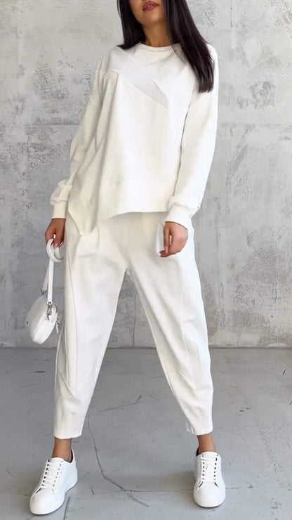 Frau in weißem, lässigem Outfit mit Pullover und Jogginghose, weiße Sneakers, kleine weiße Handtasche, vor grauer Betonwand stehend.