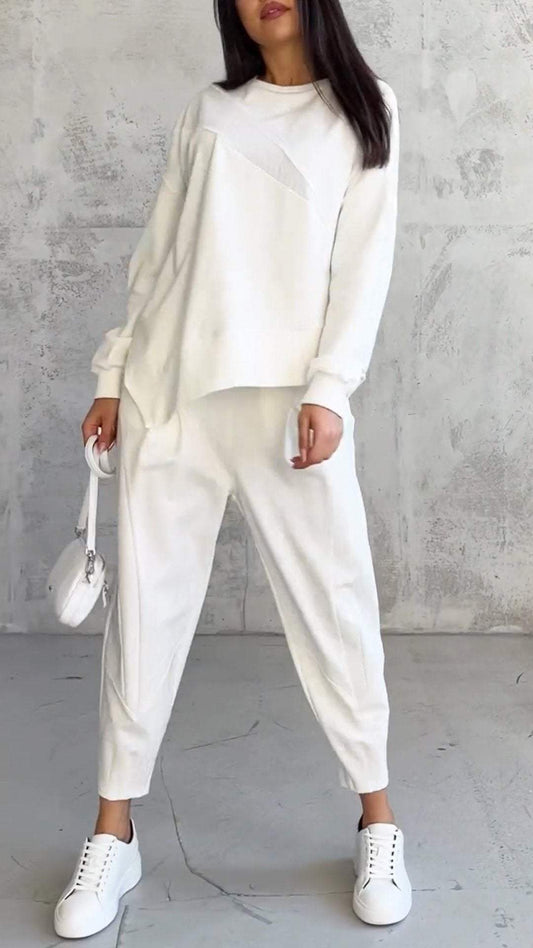 Frau in weißem, lässigem Outfit mit Pullover und Jogginghose, weiße Sneakers, kleine weiße Handtasche, vor grauer Betonwand stehend.