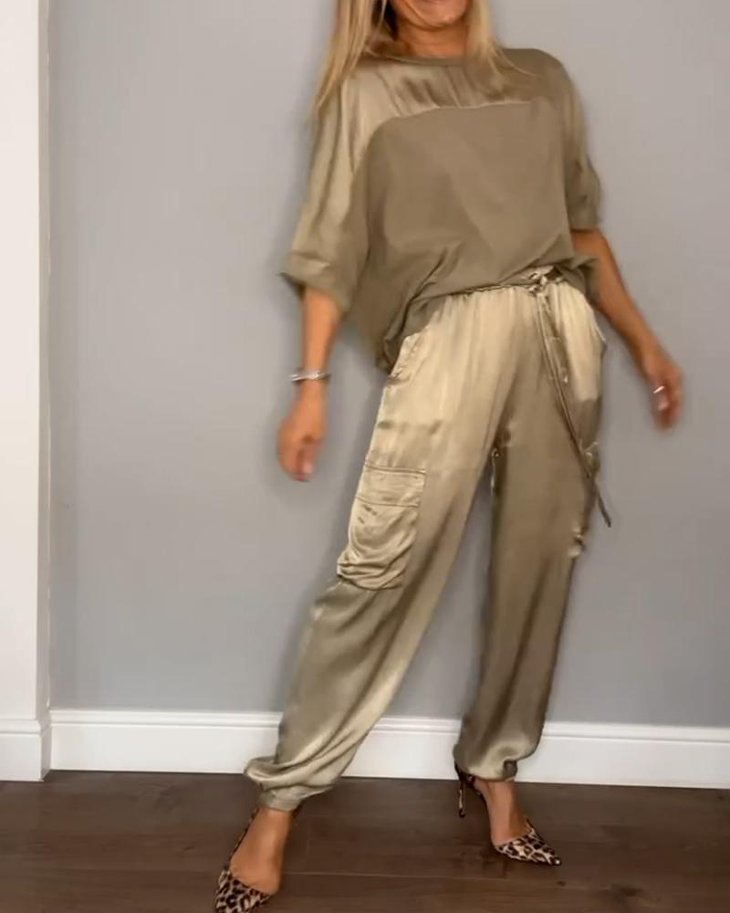 Frau in eleganter, beiger Satinbluse und Hose, posiert vor grauer Wand. Modetrend, Damenmode, Satin-Outfit, stilvolle Kleidung, Modefotografie.