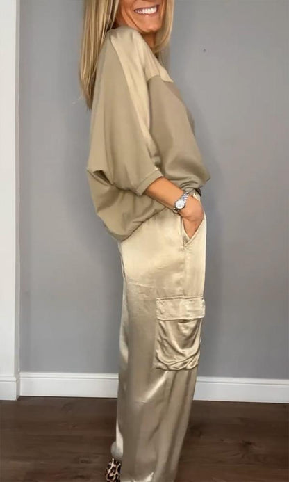 Frau in beiger Satin-Hose und lockerem Oberteil, seitlich stehend, lächelnd. Modetrend, elegante Damenbekleidung, lässiger Stil.